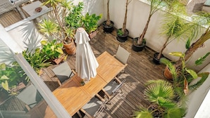 Eine Holzterrasse mit Tisch und Stühlen, umgeben von Topfpflanzen und einem weißen Sonnenschirm.
