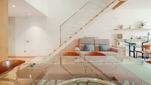 Ein modernes Interieur mit einem Glastisch, einer Couch mit Kissen, einer Treppe und einer Küchenzeile inklusive Esstisch und Stühlen.