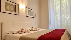 A bedroom with a large bed, a headboard, two wall-mounted light fixtures, and a window with sheer curtains.
