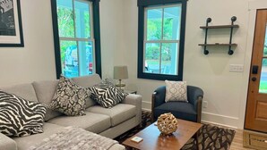 A living room with a sofa, armchair, and coffee table. There are two windows with black frames and a door with a wooden panel. - Oyster Rake Cottage, at Magnolia Court Suites (Beaufort)