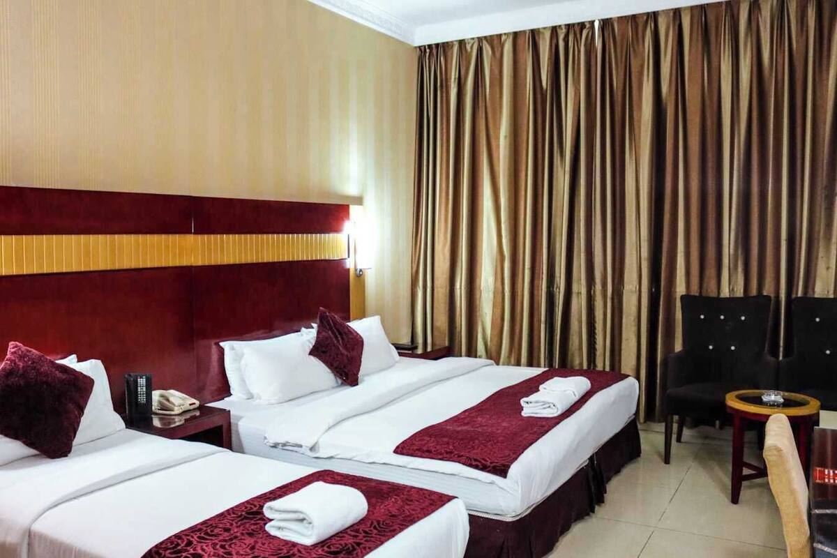 A hotel room with two beds, a wooden headboard, a curtain, a chair, and a small table.