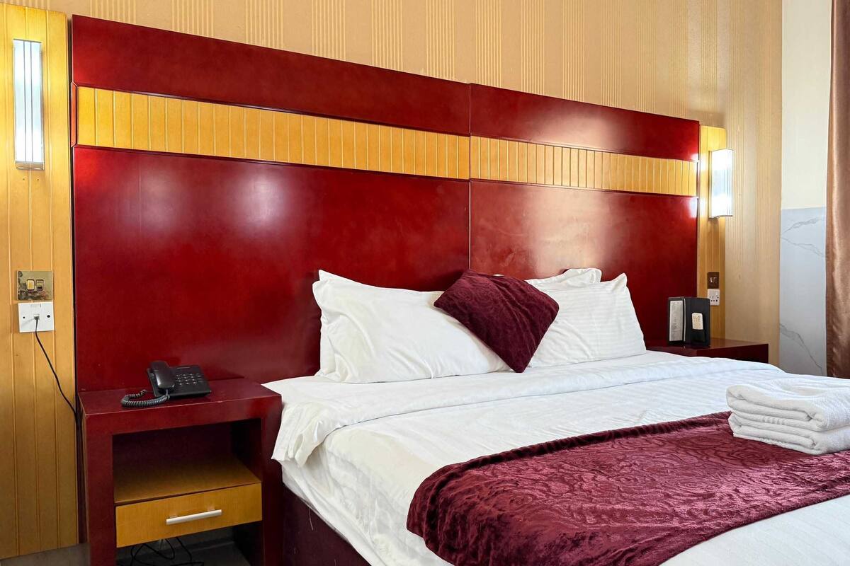 A hotel room with a large bed, a wooden nightstand with a phone, a red headboard, and a brown chair.