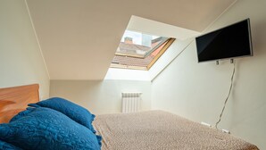 Un dormitorio con techo inclinado, tragaluz, cama con ropa de cama azul estampada de leopardo y televisor montado.