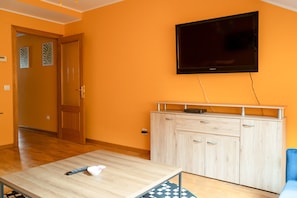 Um quarto com uma mesa de madeira, uma televisão fixada na parede, um armário claro e um sofá azul.