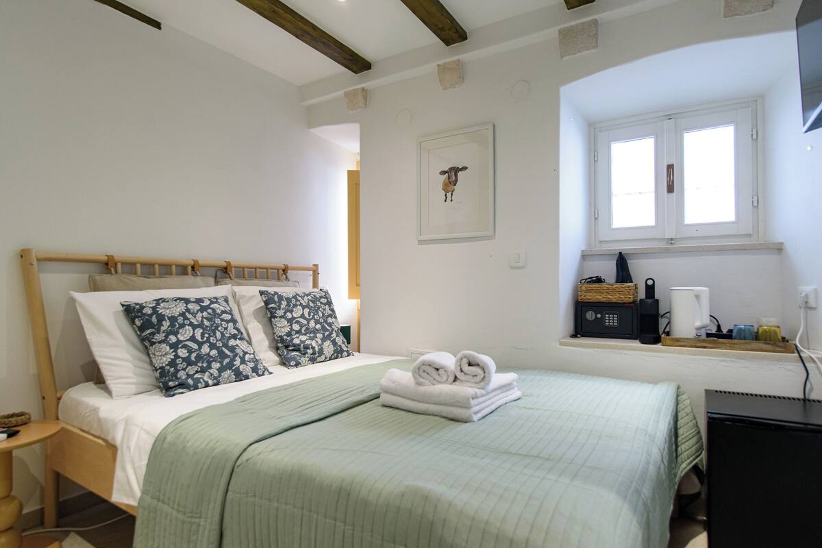 Una cama bien hecha con una colcha verde y dos almohadones decorativos, una mesita de noche con una lámpara, y una ventana con vistas al exterior.