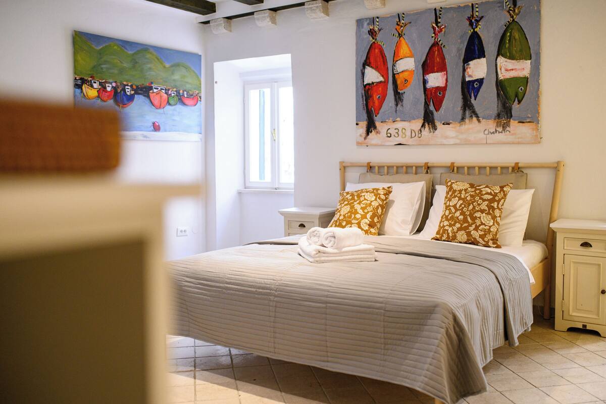 Una cama bien hecha con cubrecama a rayas y dos almohadas, un cuadro de peces de colores en la pared, y una ventana que proporciona luz natural.