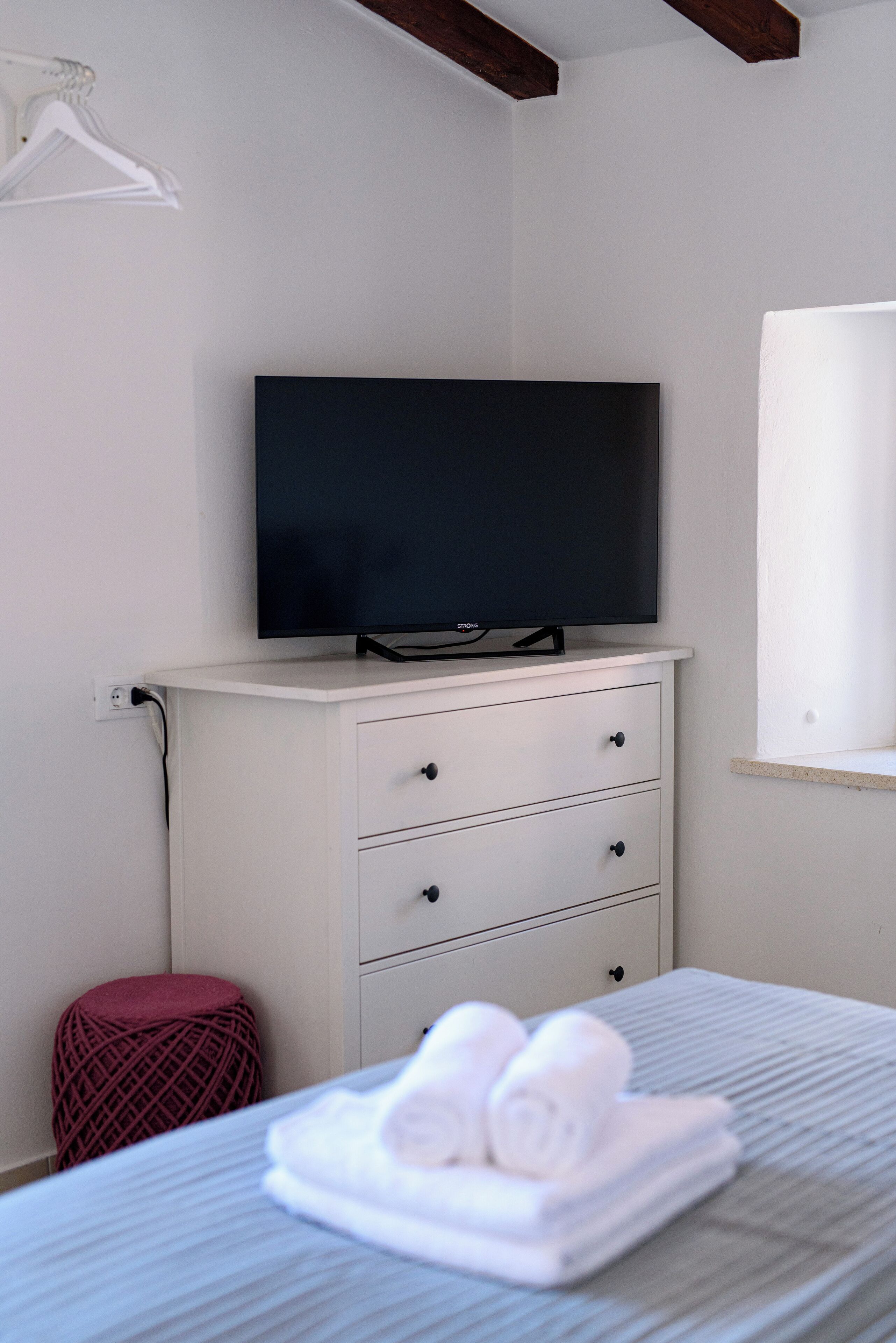 Une chambre avec un lit, un téléviseur posé sur une commode, un pouf rouge et une fenêtre.