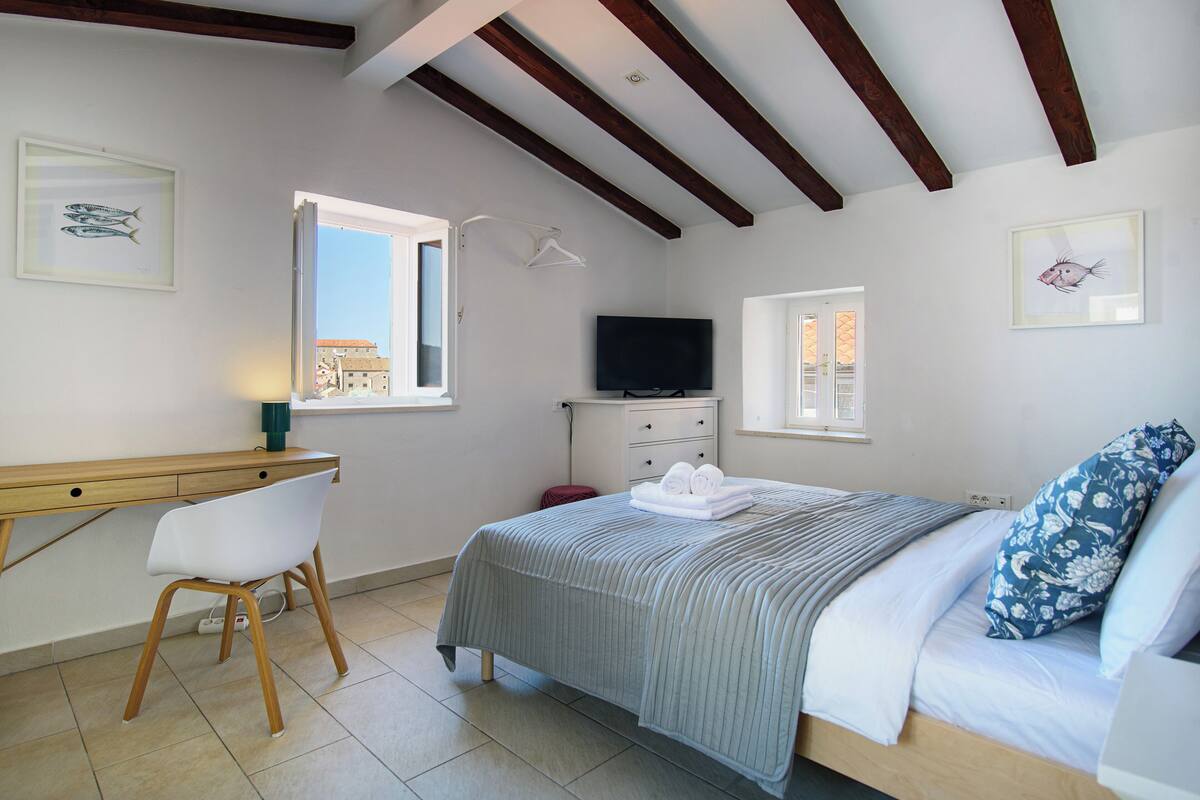 Un dormitorio con cama, escritorio, silla, televisor y obras de arte en las paredes.