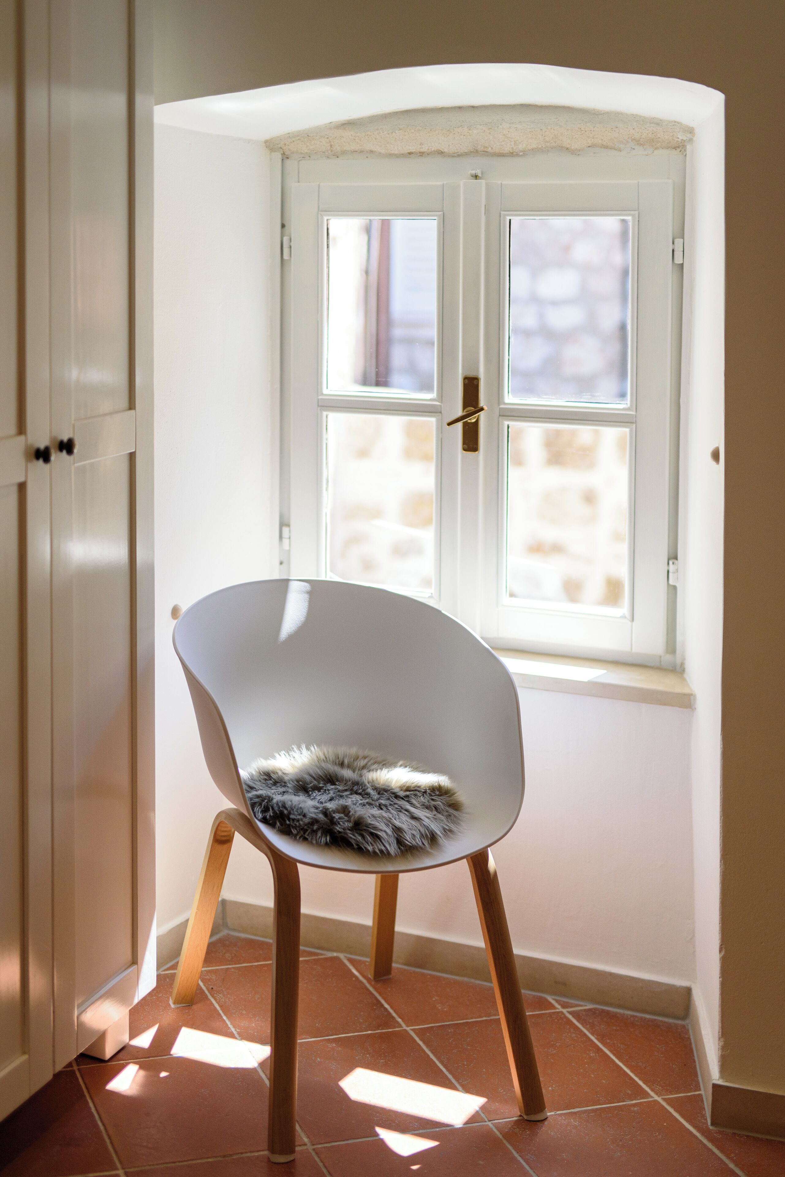 A white chair with a fluffy cushion in a corner of a room with a white door and a window with a view of a stone wall.