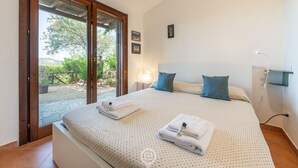 Une chambre avec un grand lit, une porte-fenêtre coulissante donnant sur un jardin, une table de chevet avec une lampe et une étagère fixée au mur.