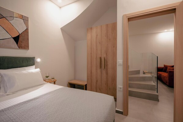 Une chambre moderne avec un grand lit, une armoire en bois, une petite table de chevet et un escalier en verre menant à un espace de vie agrémenté d’un canapé rouge.