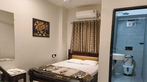 A hotel room with a bed, a bedside table with a lamp, a mirror, a painting on the wall, and a bathroom visible through a glass door. - Saryu Homestay (Ujjain)