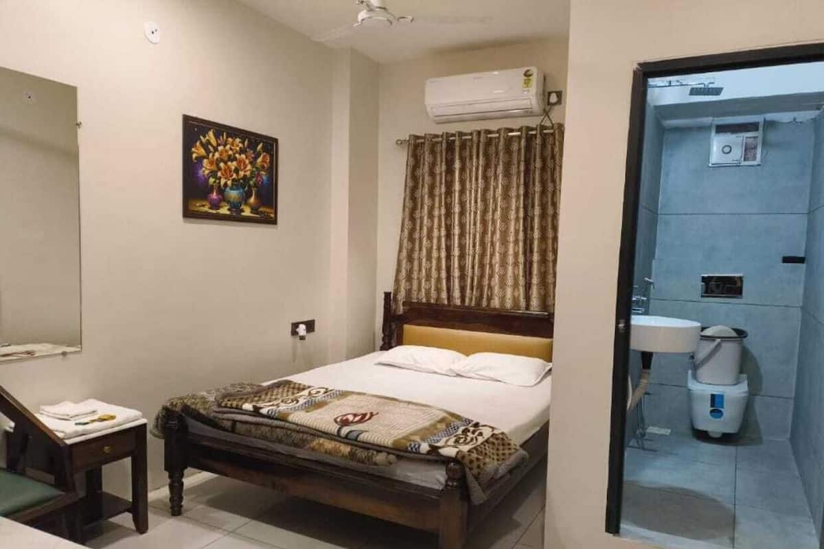 A hotel room with a bed, a bedside table with a lamp, a mirror, a painting on the wall, and a bathroom visible through a glass door.