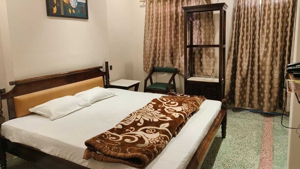 A hotel room with a bed, a chair, a small table, an air conditioning unit, and curtains. - Saryu Homestay (Ujjain)