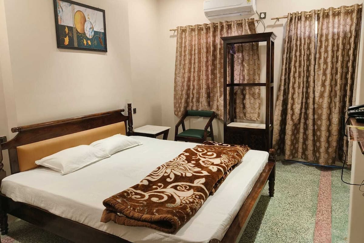 A hotel room with a bed, a chair, a small table, an air conditioning unit, and curtains.