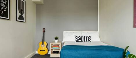 A bedroom with a bed, a guitar, a small shelf with books, and a plant.