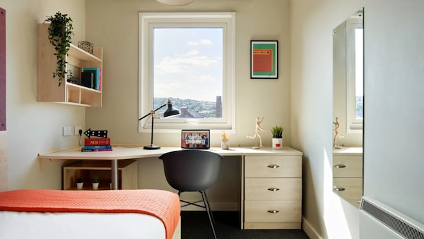 Ein kompaktes Hotelzimmer mit Bett, Schreibtisch mit Computer, Stuhl, Fenster mit Stadtblick und einer kleinen Pflanze.