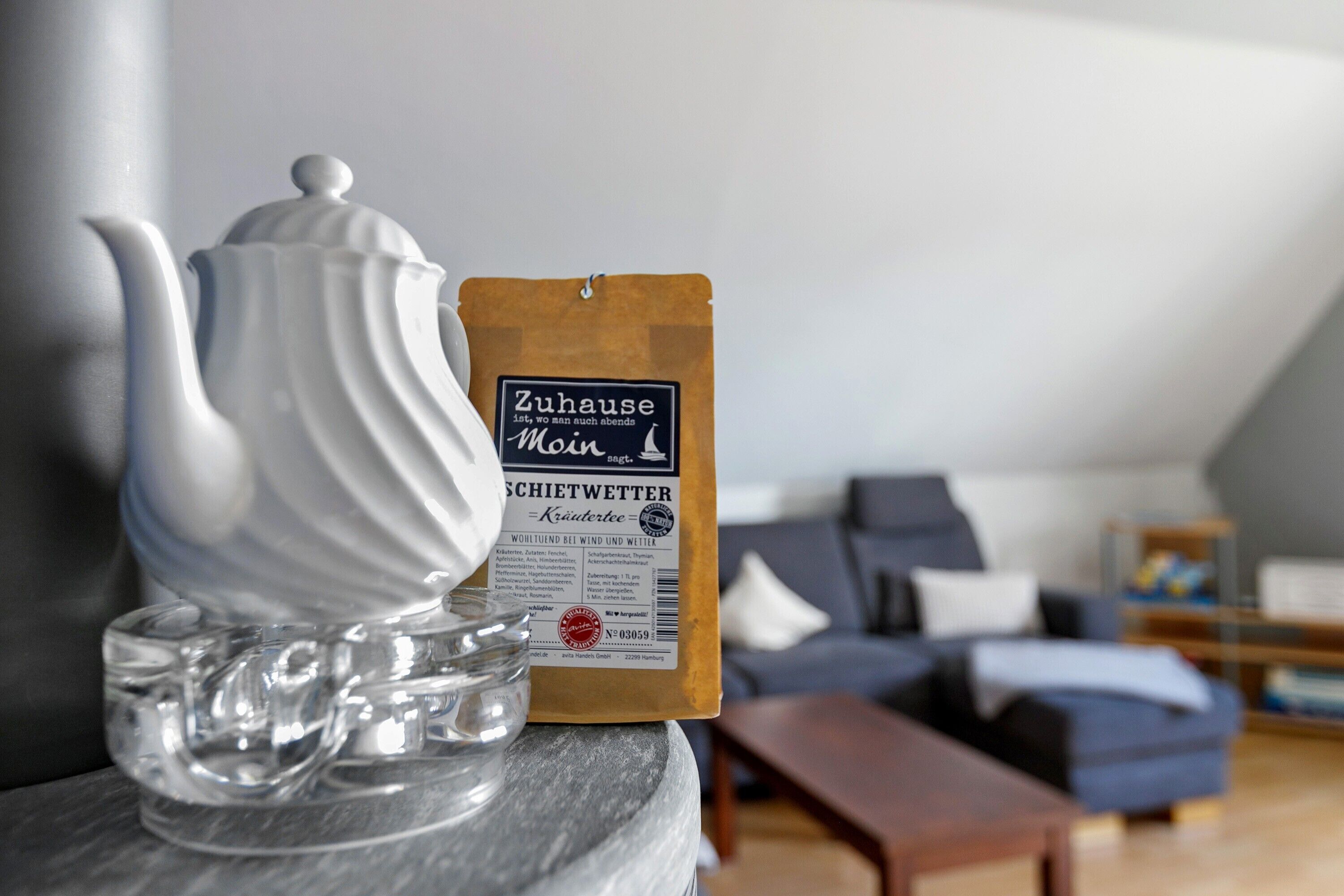 A teapot, a glass, and a coffee bag on a marble surface in a living room with a sofa and a small table.