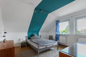Une chambre avec un plafond en pente, un lit recouvert d’une literie à motifs, une table de chevet avec une lampe, une chaise et une fenêtre donnant sur un paysage verdoyant.