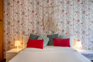 A bedroom with a bed, pillows, a hanging macrame, and a wall with a tree and bird pattern.