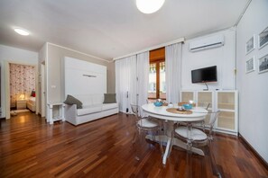 Una sala de estar moderna con un sofá blanco, una mesa de comedor con sillas, un televisor montado en la pared y un equipo de aire acondicionado.