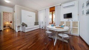 A modern living room with a white sofa, a dining table with chairs, a TV mounted on the wall, and an air conditioning unit. - GuestHost - Cozy 53 sqm apartment, perfect for four people, located on the first floor of a building with an elevator (NOT accessible to disabled guests).The property is located in Grado, known as the Island of the Sun, a beautiful lagoon town famous for (Grago GO)