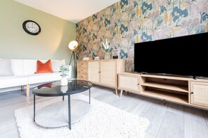 Un salon comprenant un canapé, une table basse, un téléviseur posé sur un meuble en bois, une horloge murale et un papier peint à motifs floraux.