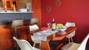 Un coin repas avec une table en bois, des chaises blanches, un mur rouge et une cuisine en arrière-plan.