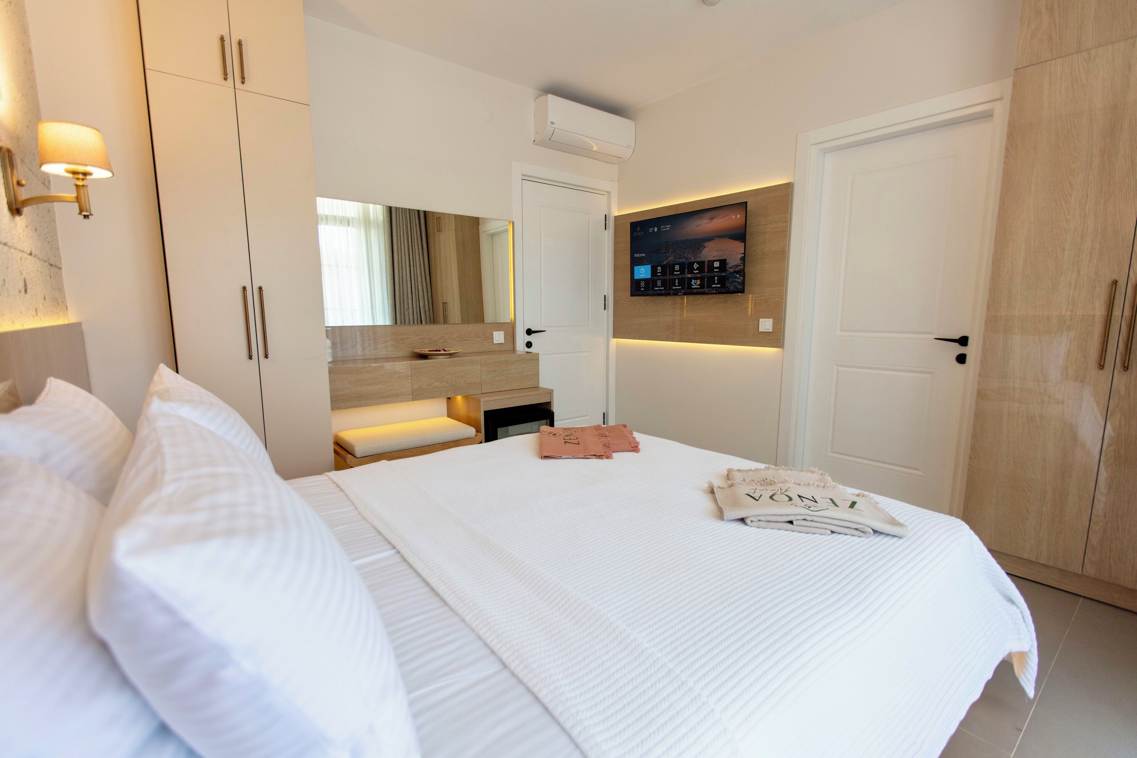 A hotel room with a large bed, a vanity with a mirror, a flat-screen TV mounted on the wall, and a wardrobe.