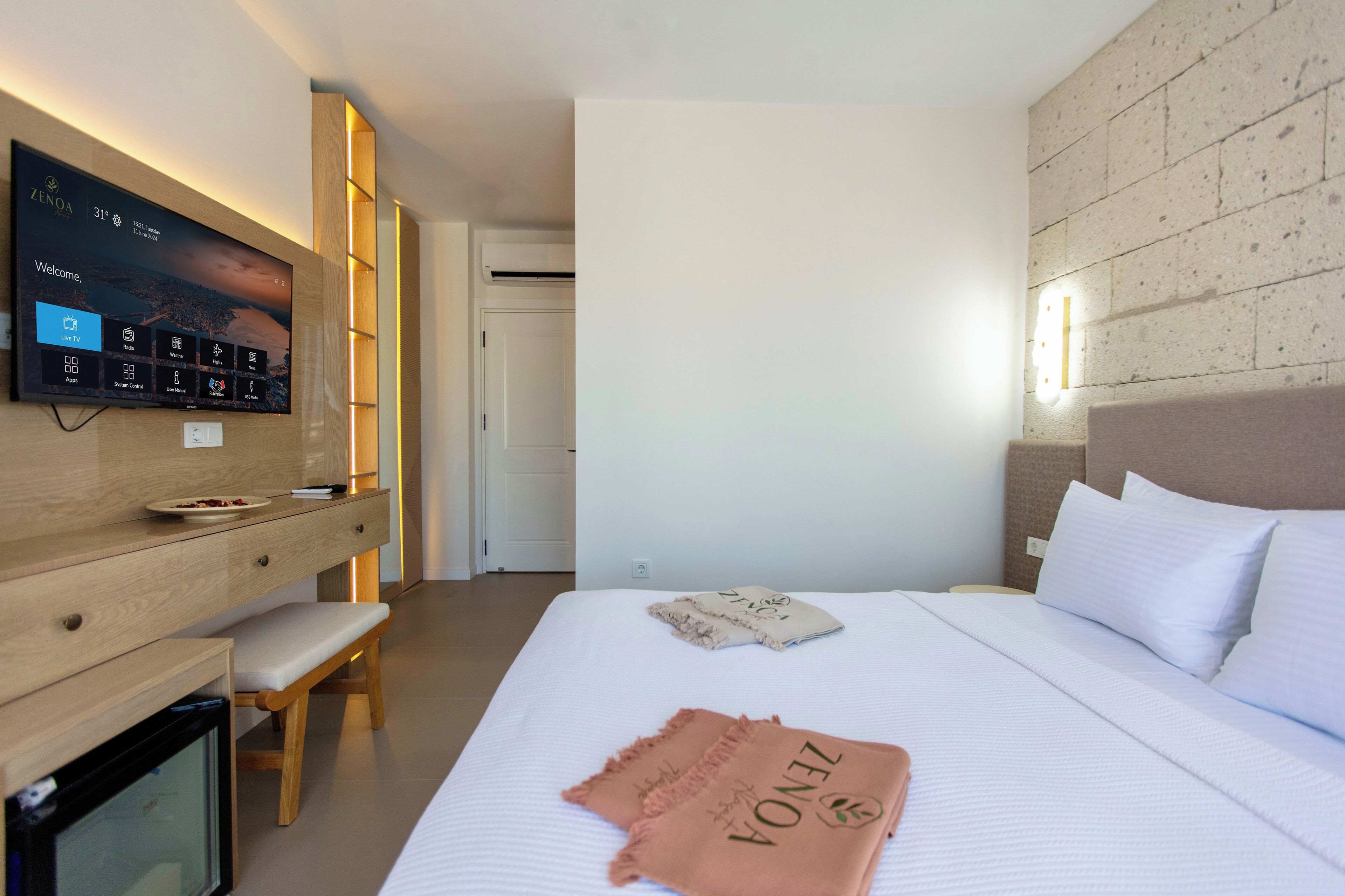 Une chambre d’hôtel moderne dotée d’un grand téléviseur à écran plat fixé au mur, d’un bureau en bois avec une banquette, d’un lit avec une literie blanche et une couverture ZINQ, et d’un mur décoratif à l’aspect pierre.