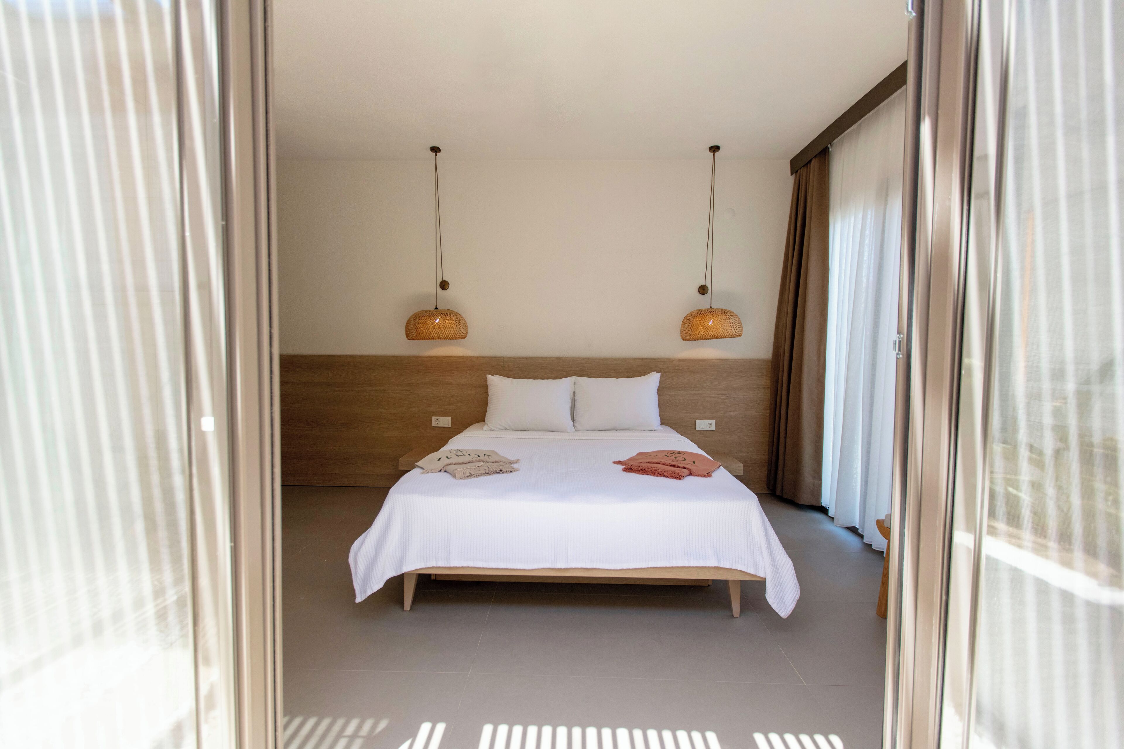 Une chambre moderne avec une tête de lit en bois, un grand lit recouvert de linge de lit blanc, deux lampes suspendues et une vue sur l’extérieur grâce à de grandes fenêtres.