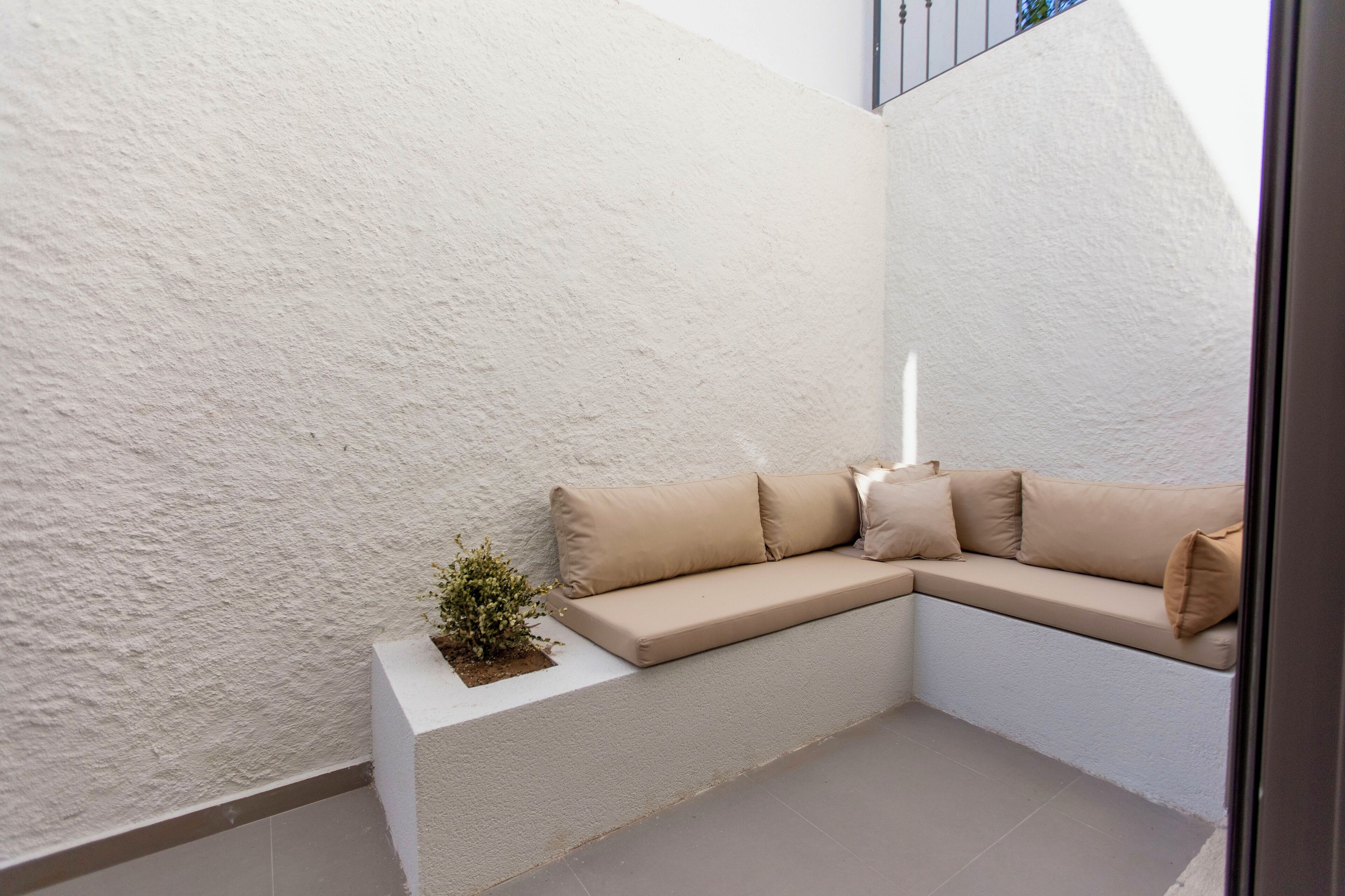 A corner seating area with a beige sofa and cushions, a small plant on a white planter, and a white textured wall.