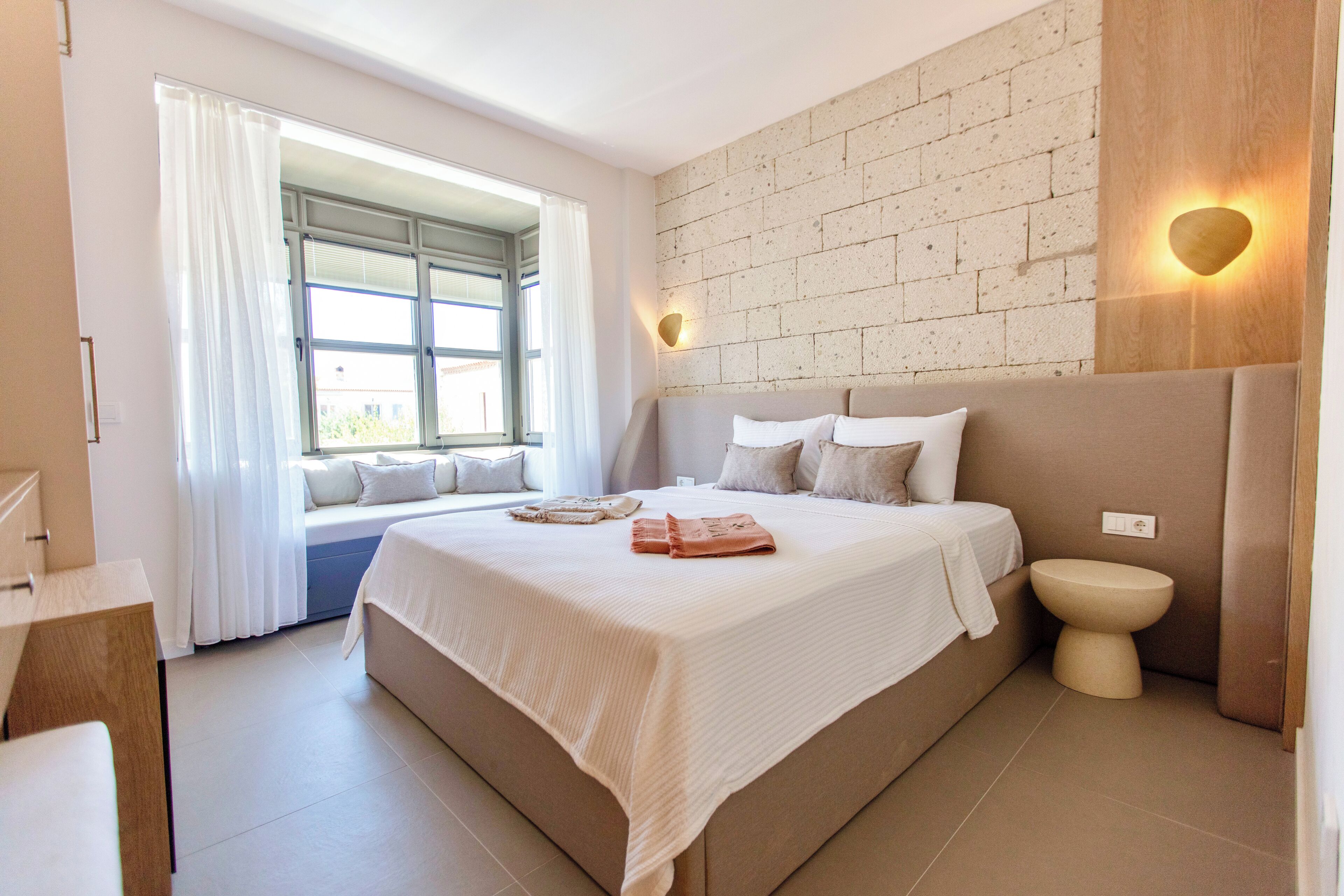 A modern bedroom with a large bed, a bedside table, a window with curtains, a brick wall, and a wall-mounted light fixture.