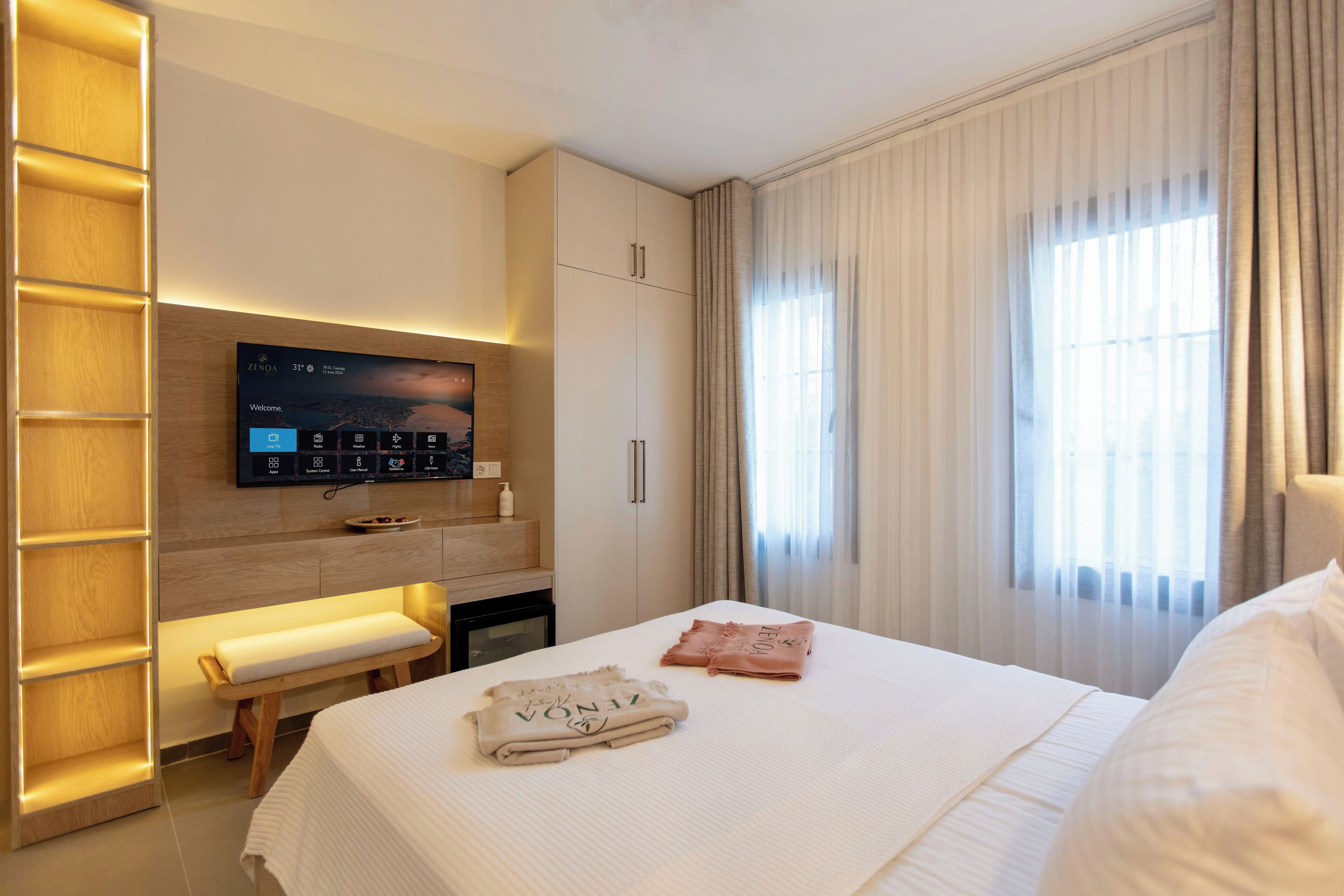 A hotel room with a bed, a TV mounted on the wall, a wooden bench, and a window with curtains.