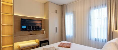 A hotel room with a bed, a TV mounted on the wall, a wooden bench, and a window with curtains.