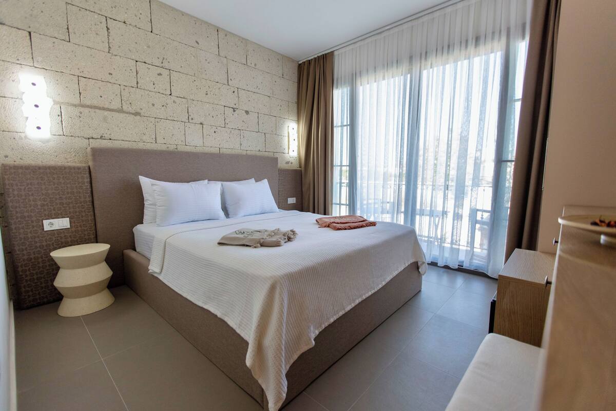A hotel room with a large bed, a bedside table, a wall-mounted light, and a window with curtains.