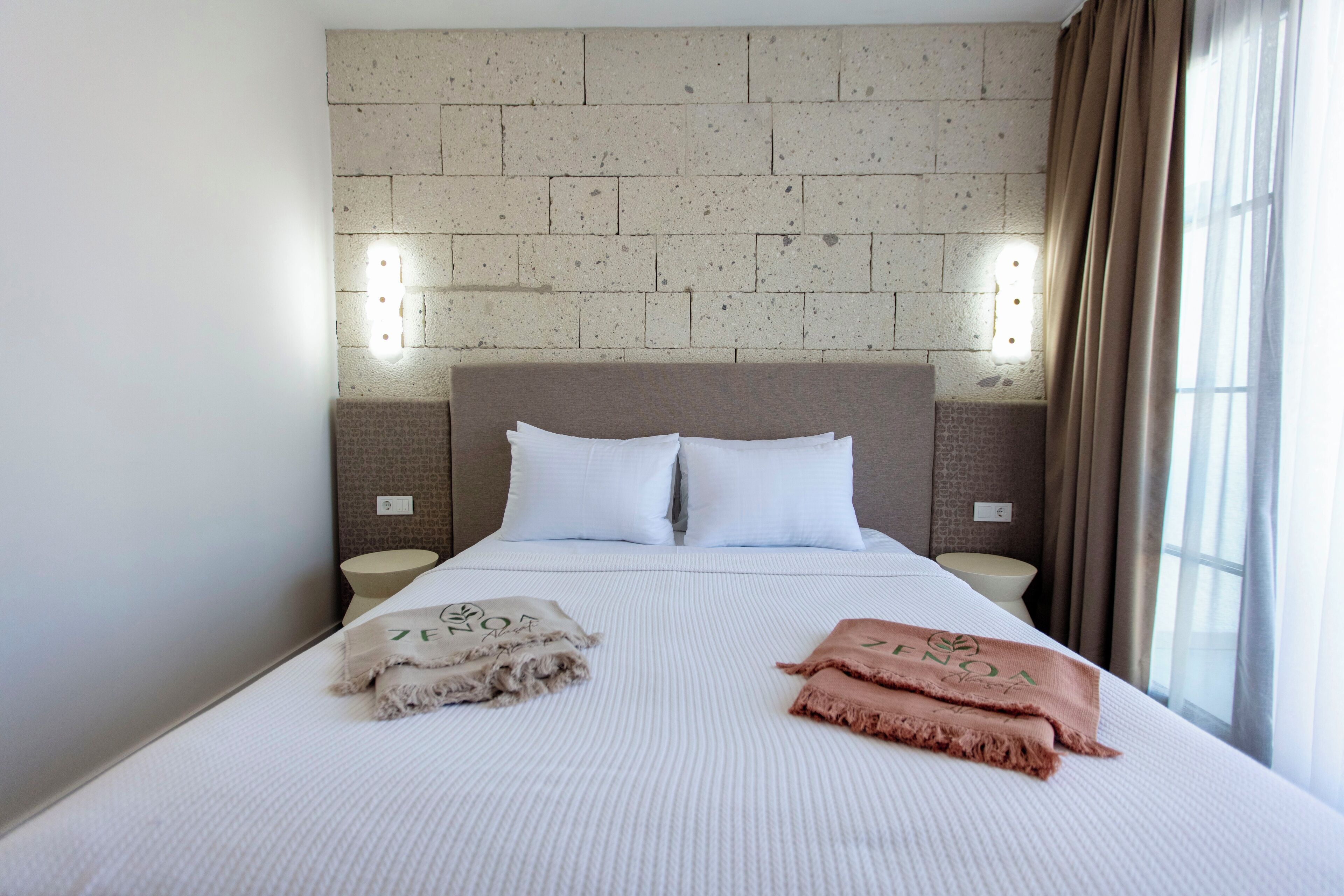 A neatly made bed with two pillows and two blankets, a brick-patterned headboard, bedside lamps, and a window with curtains.