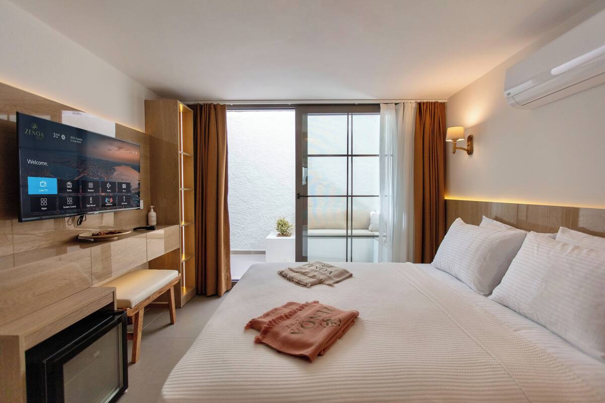 A hotel room with a bed, a TV mounted on the wall, a bedside table, a bench, and a view of the outdoors.