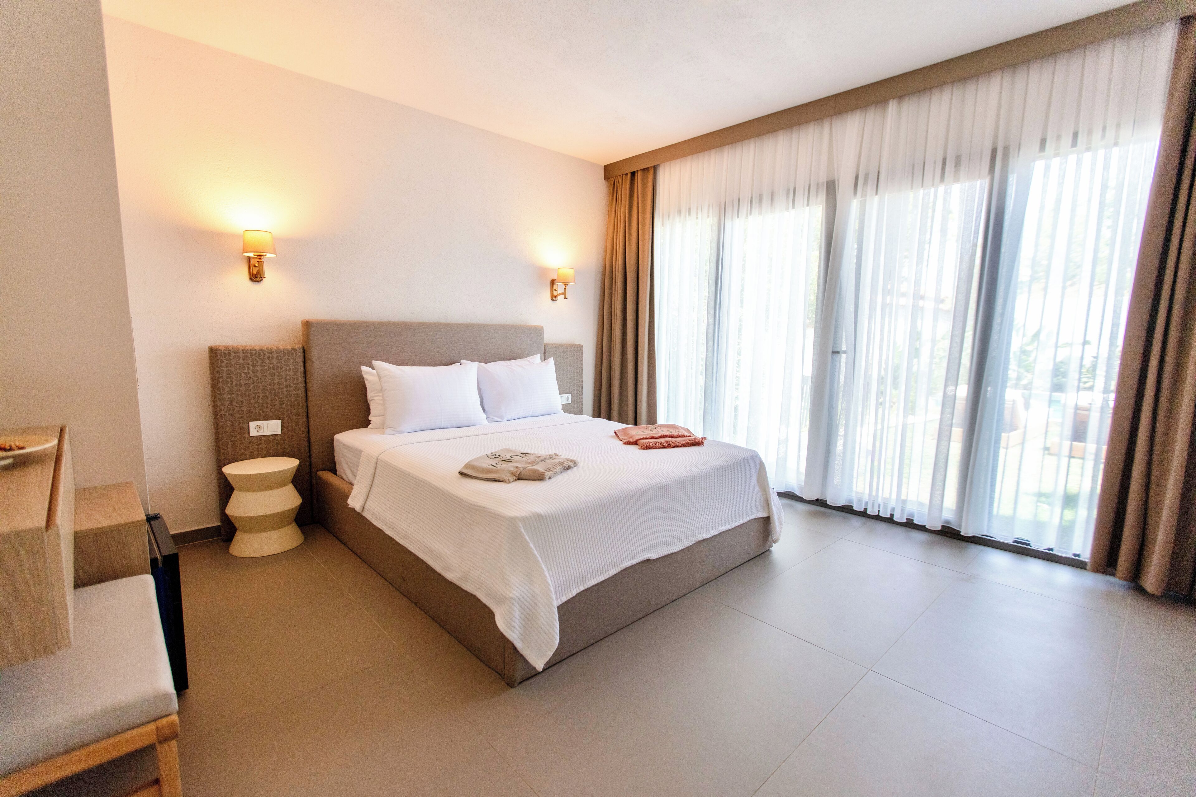 Habitación de hotel con una cama grande, dos lámparas de noche, una silla de madera, una mesita redonda y un ventanal con cortinas.