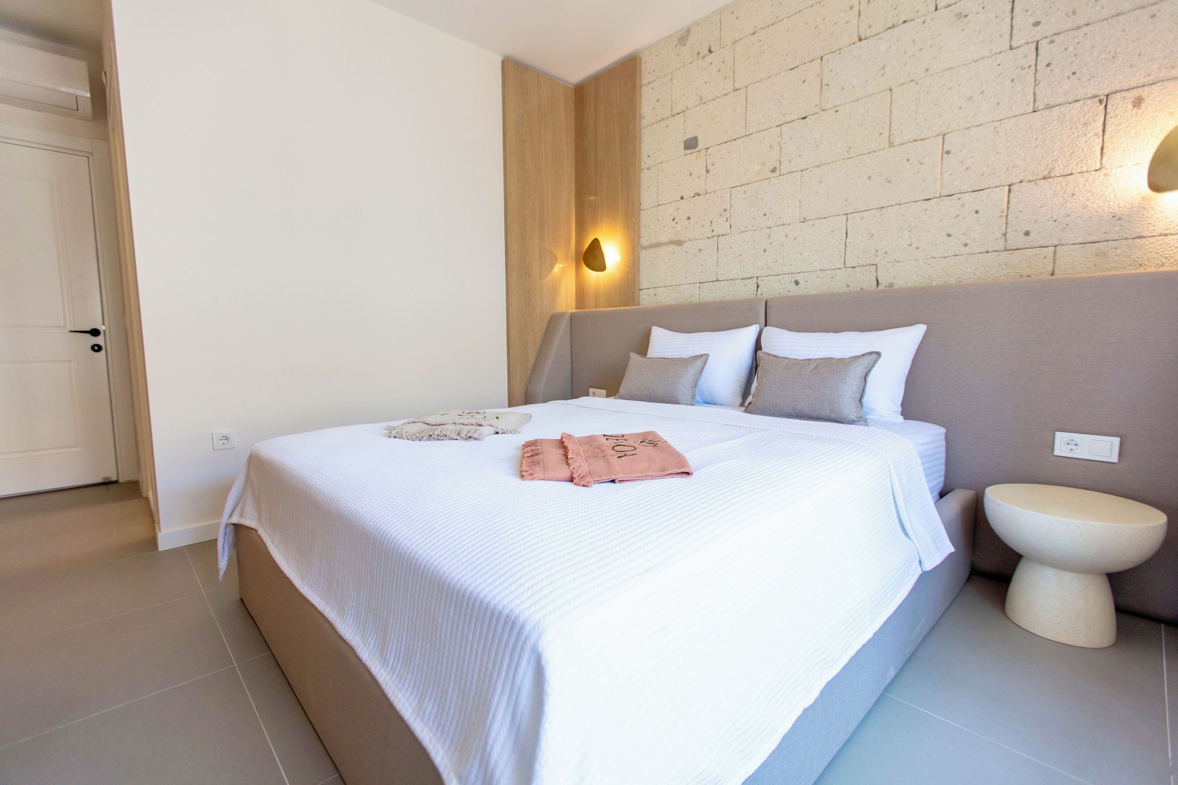 Cama bem arrumada com colcha branca e bege, toalha rosa e cabeceira em frente a uma parede de tijolos com luminária.