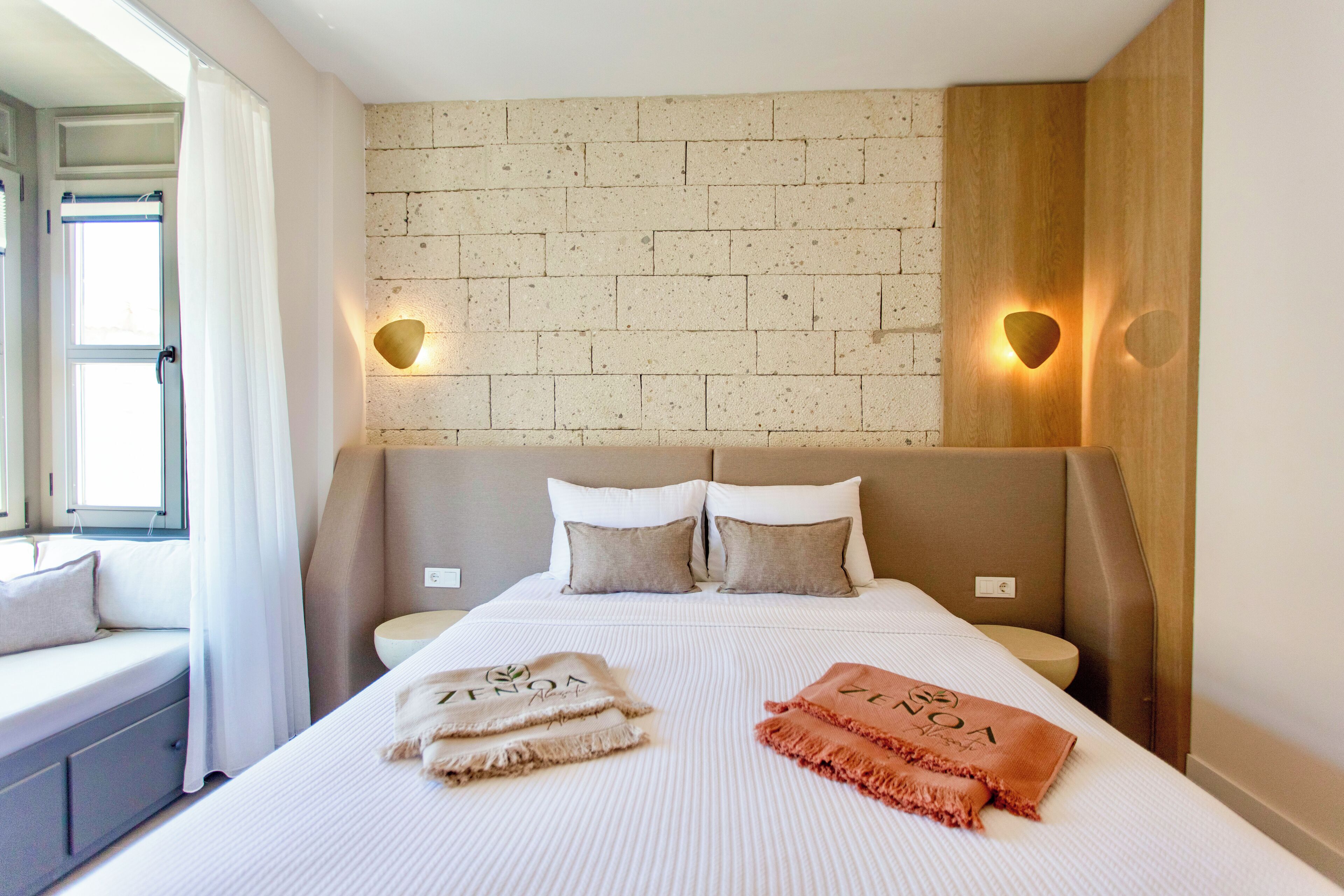 A bedroom with a brick wall, two bedside tables, a bed with white bedding and two pillows, and two wall-mounted lights.