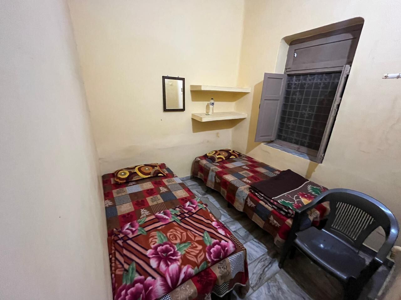 A small room with two beds, a chair, a shelf with a bottle, and a window with a metal grill.