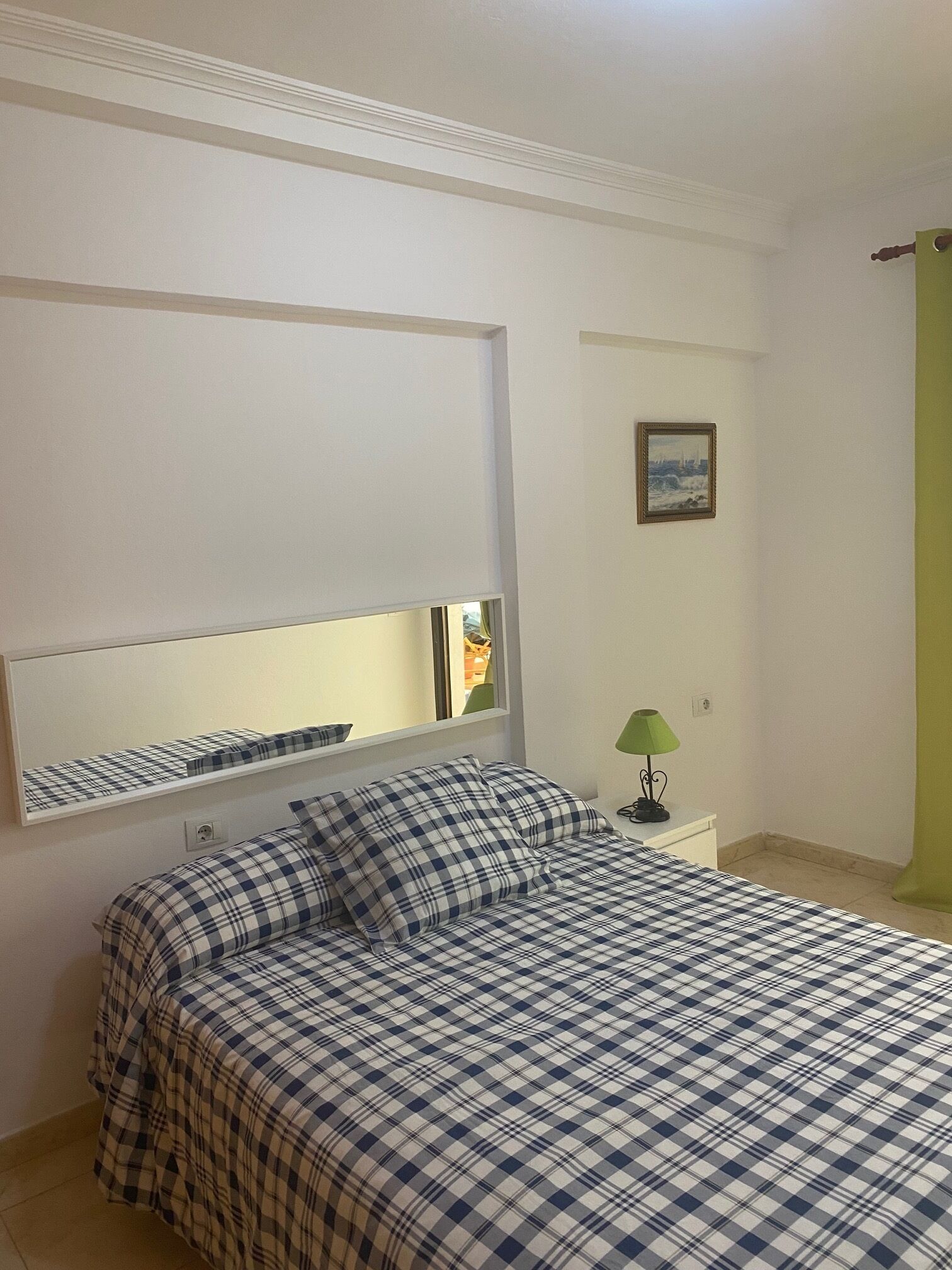 Een slaapkamer met een bed, een nachtkastje, een lamp, een ingelijst beeld en een raam met uitzicht op een stadsbeeld.