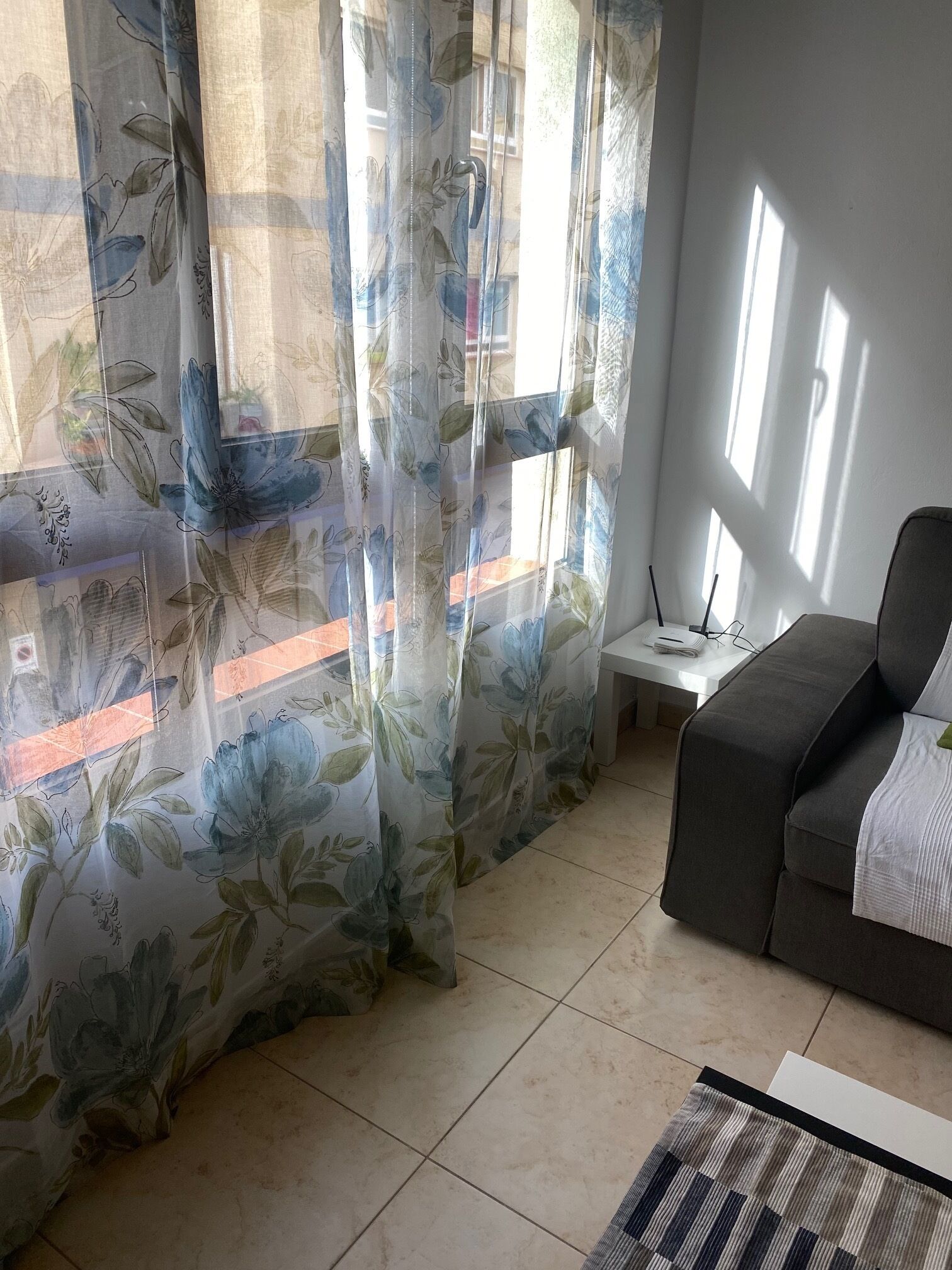 Ein Zimmer mit einem grauen Sofa, einem kleinen weißen Tisch mit einem Laptop und einem Fenster mit floralen Vorhängen.
