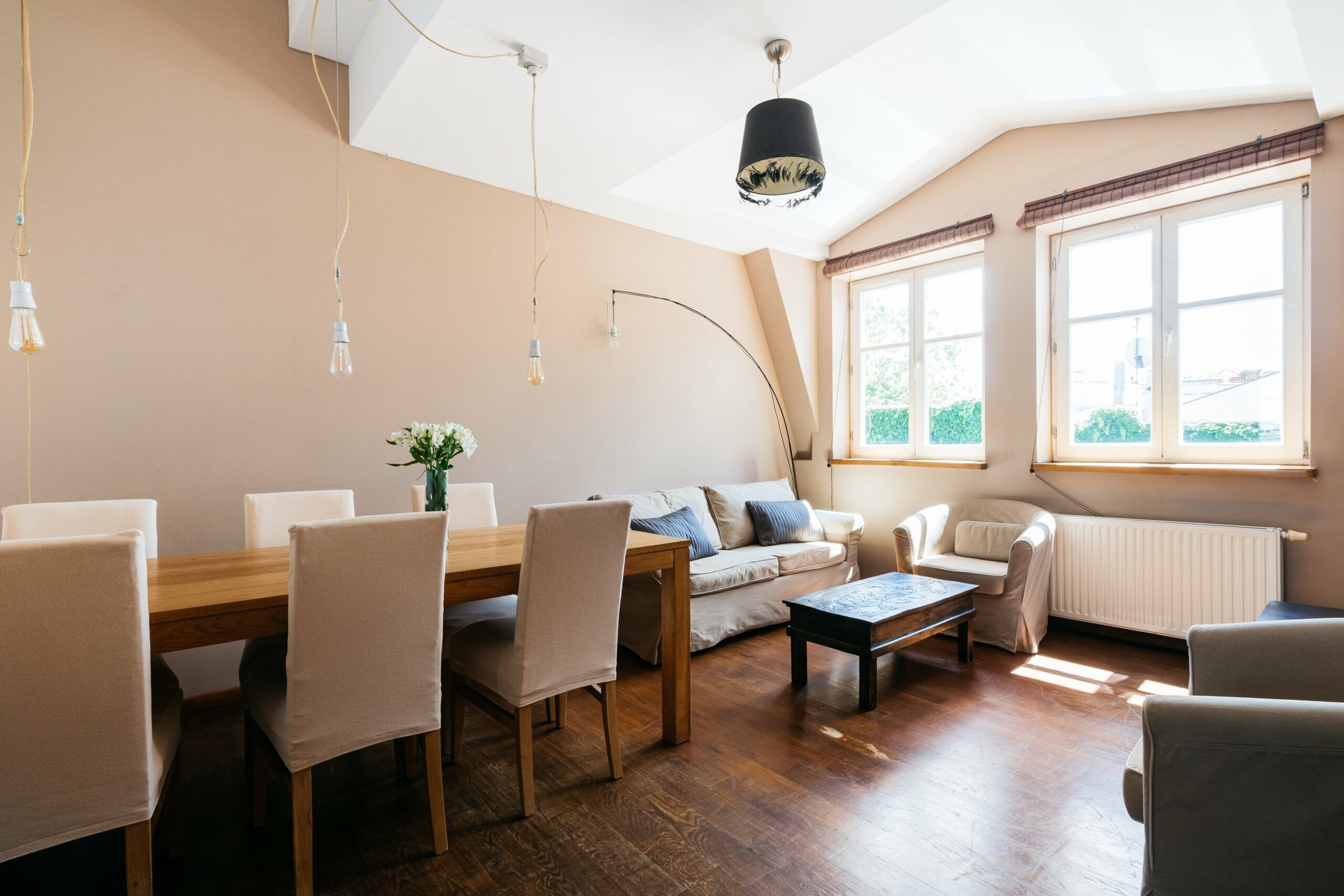 Una sala de estar moderna con una mesa de comedor de madera, sillas color beige, un sofá y una mesa de centro. Hay ventanas amplias con cortinas, un radiador y una lámpara en el techo.