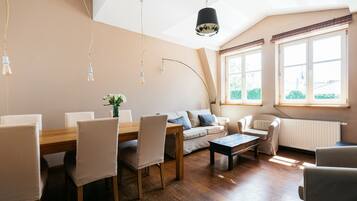 Una sala de estar moderna con una mesa de comedor de madera, sillas color beige, un sofá y una mesa de centro. Hay ventanas amplias con cortinas, un radiador y una lámpara en el techo.