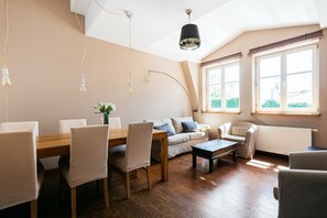 Uma sala de estar moderna com mesa de jantar de madeira, cadeiras bege, um sofá e uma mesa de centro. Há janelas amplas com cortinas, um radiador e um lustre.