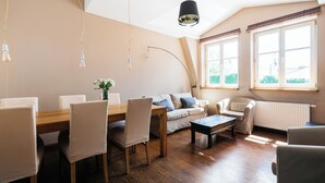 Een ruime woonkamer met een houten eettafel, beige stoelen, een bank en een salontafel. Er zijn grote ramen met gordijnen, een radiator en hangende lampen.
