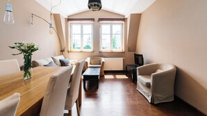 Een ruime woonkamer met een houten eettafel, twee fauteuils, een bank en een televisie. Grote ramen zorgen voor veel natuurlijk licht.