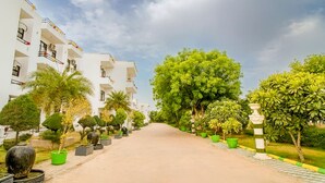 A pathway leading to a white building with balconies, surrounded by lush greenery and trees.